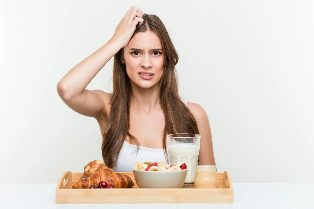 Sieviete apjukusi pie brokastu galda — piena glāze, pārslas un maize; izteikts apjukums par uzturu, ja trūkst olbaltumvielas.