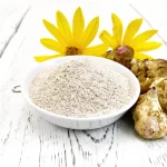 Topinambūru pulveris baltā bļodā ar svaigiem topinambūriem un ziediem uz balta galda